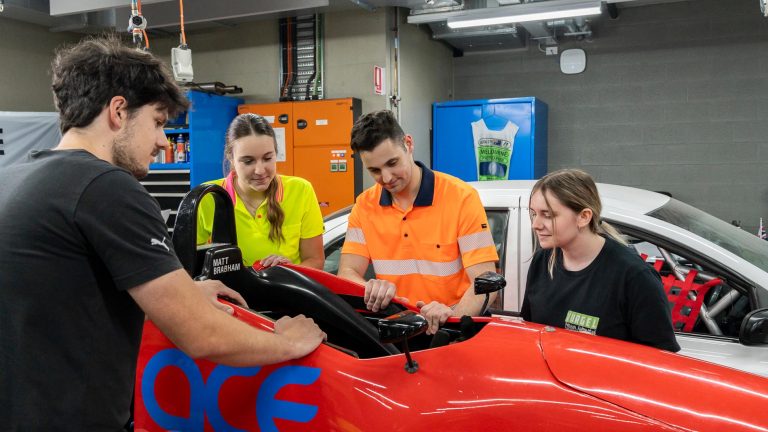 Four students in high-visibility and black shirts are gathered around the open cockpit of a red race car, likely a formula or open-wheel vehicle, in a workshop setting.