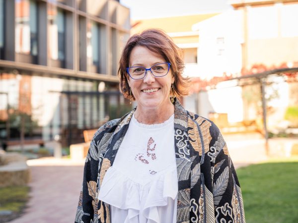 A close-up outdoor portrait of a smiling woman. She has shoulder-length reddish-brown hair and is wearing blue-framed glasses. She is wearing a white blouse with a patterned cutout on the front and a dark jacket with an orange and white batik-style pattern. The background is a modern building exterior and a courtyard with greenery, slightly out of focus.